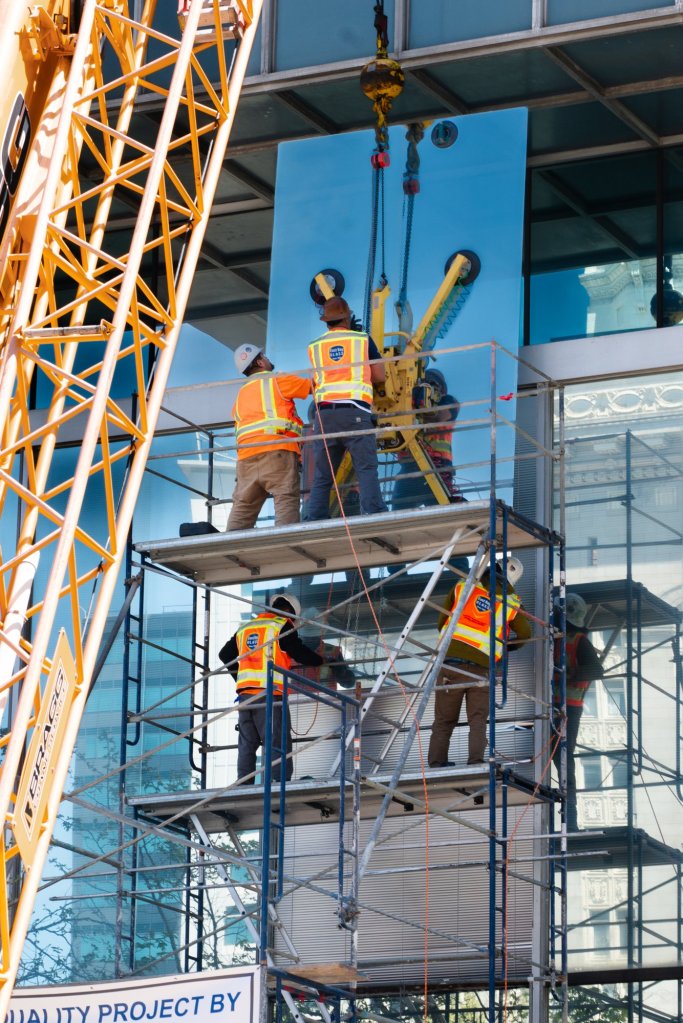 East Bay Glass glaziers on scaffolding setting a large curtain wall glass panel with a crane and vacuum lifter in downtown Oakland