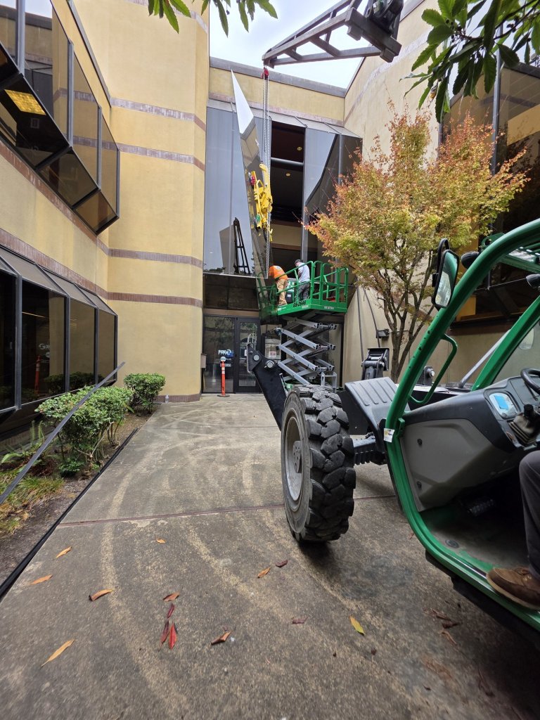 Telehandler lifting a tall glass panel in a tight courtyard at the KTVU Fox 2 building in Oakland