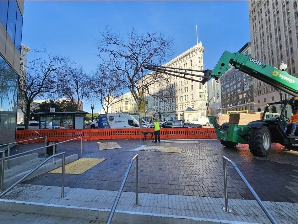 Telehandler with a quad-cup vacuum lifter moving storefront glass across a downtown Oakland plaza